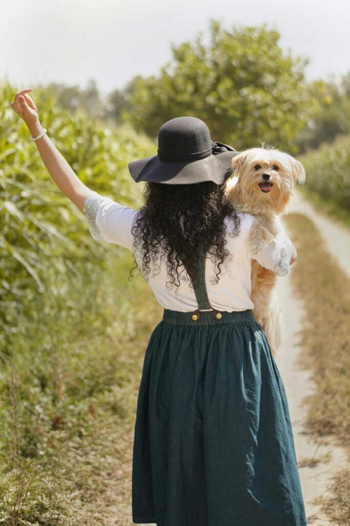 Woman in vintage dress walking on dirt road holding her dog, enjoying nature.